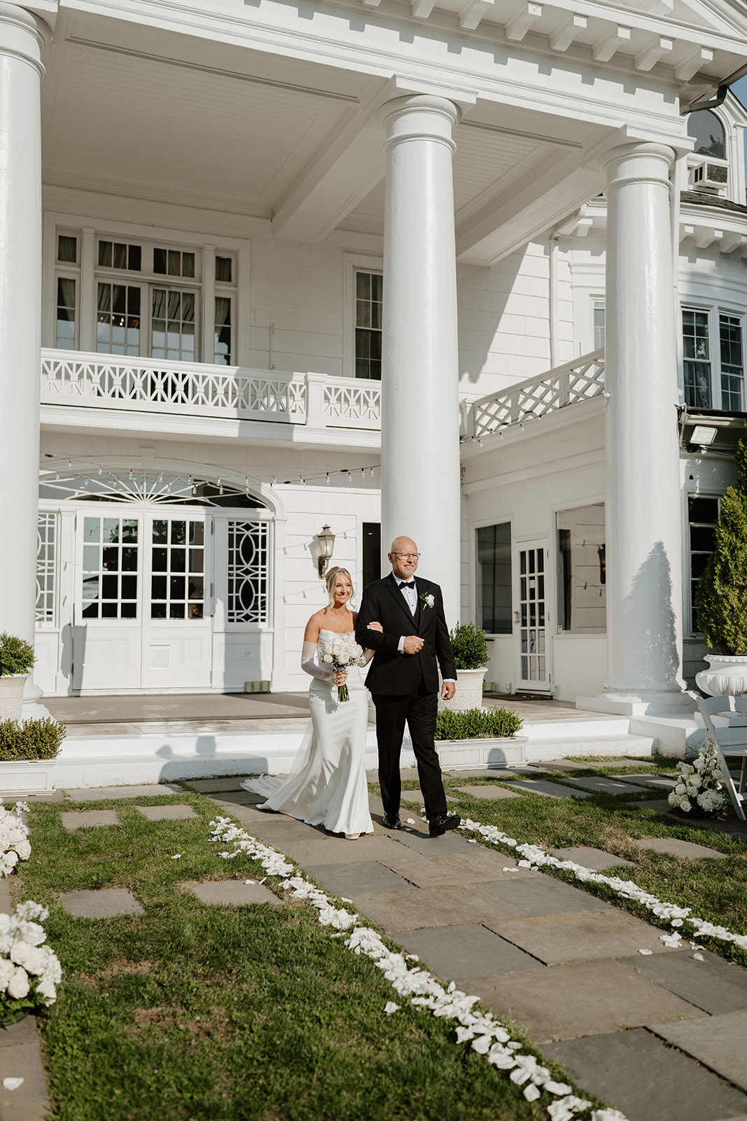 Bride walks down the outdoor aisle arm-in-arm with her father, smiling in front of the grand manor.